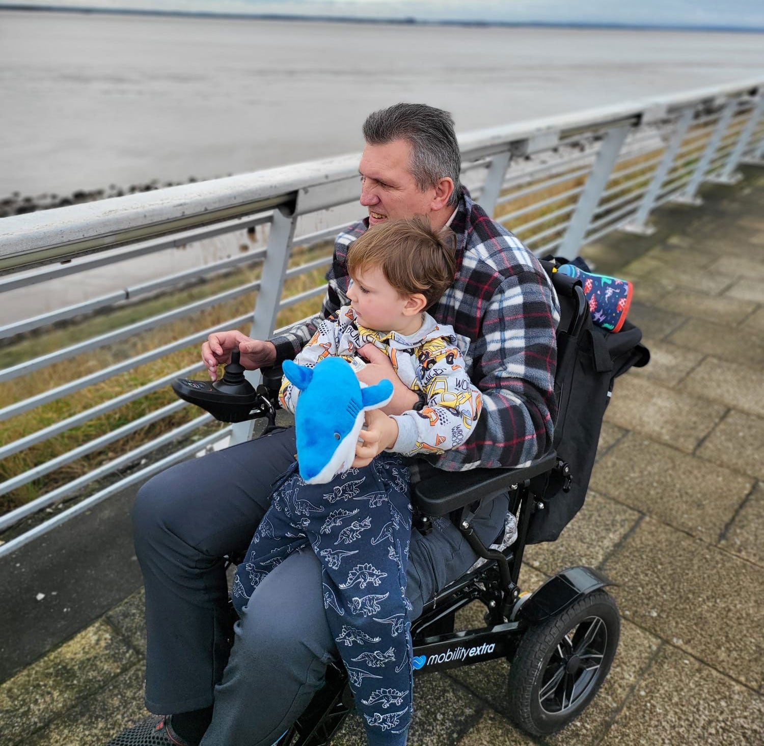 Man with a child on his lap by the sea sitting in a Mobility Extra electric wheelchair