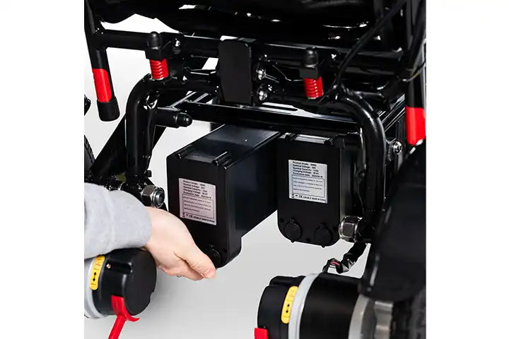 A close-up of a person's hand connecting a black battery to an electric wheelchair frame with red and black components.